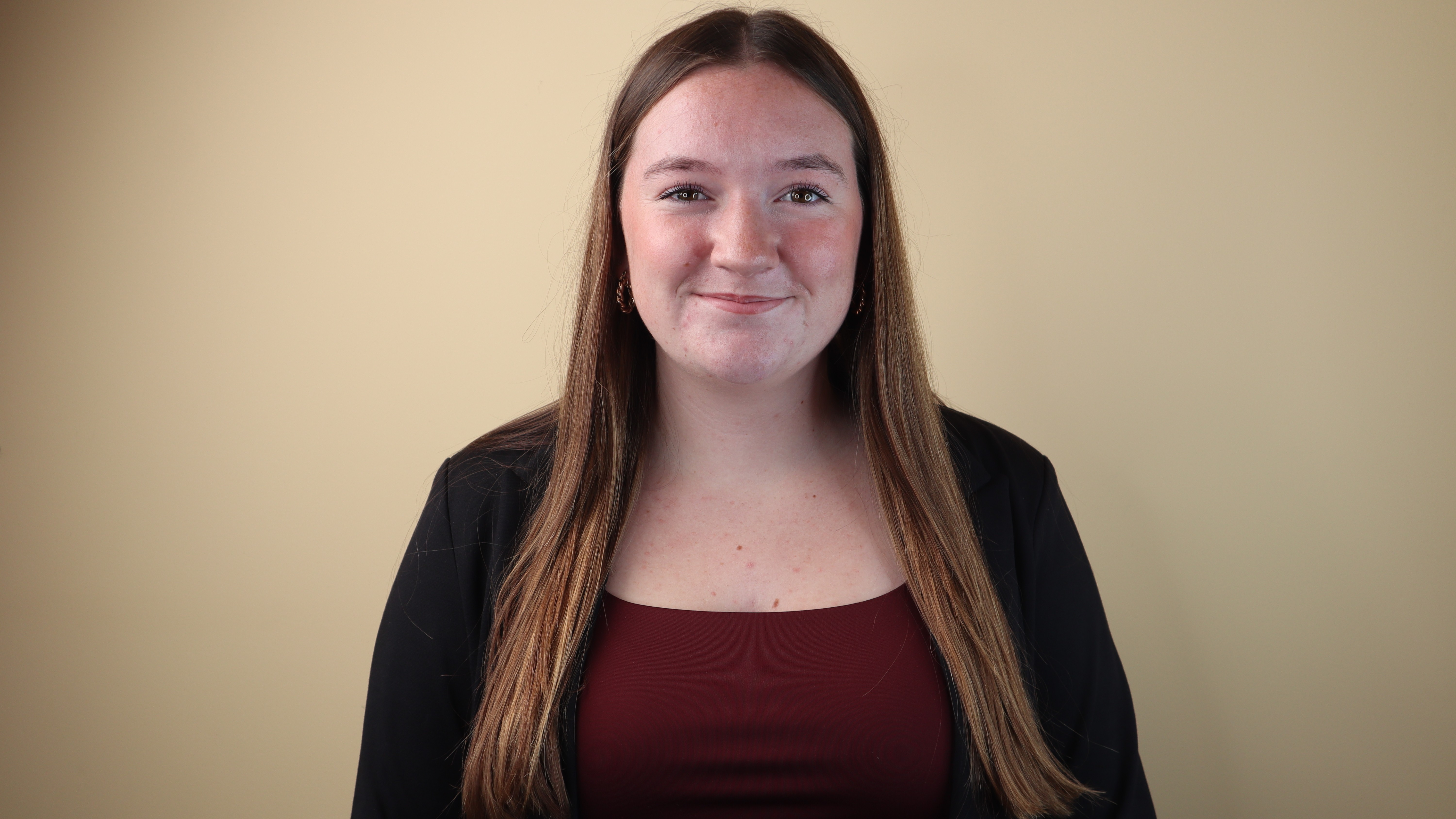 Jo Marsh smiles in front of a beige background while wearing a black blazer over a maroon shirt. Jo Marsh smiles in front of a beige background while wearing a black blazer over a maroon shirt.